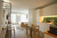 Modern studio apartment with integrated kitchen, glass dining table, and bed separated by vertical slats, showcasing a minimalist space-saving design.