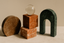 A minimalist arrangement of marble shapes in brown, orange, peach, green, and grey tones, with a glass sphere on top, set against a beige textured wall.