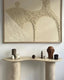 A travertine console table with a curved design, decorated with sculptures, a candle, and a vase, set against a textured abstract artwork in neutral tones.