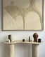 A travertine console table with a curved design, decorated with sculptures, a candle, and a vase, set against a textured abstract artwork in neutral tones.